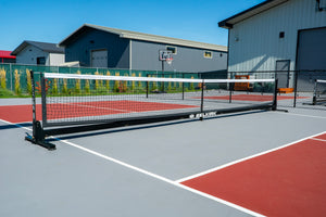 Outdoor pickleball court with SELKIRK-branded net across center, red and gray court surface and buildings in background.