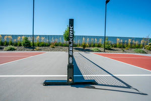 Portable volleyball net post with SELKIRK logo centered on an outdoor court, net shadow extending to the right