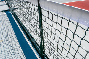 Close-up of tennis net, post and shadow cast on blue and red court surface.
