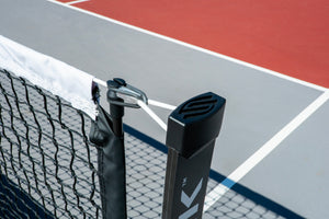 Close-up of a tennis net post with tensioning clamp and strap; white court lines on red and gray hardcourt.