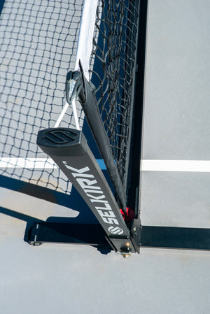 Tennis net post labeled SELKIRK with tension cord and gray court line