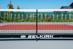 Close-up of pickleball net base with SELKIRK logo; red and gray court and fence in background.
