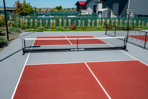 Outdoor red-and-gray pickleball courts with a SELKIRK net, fence, landscaping and a basketball hoop in the background.