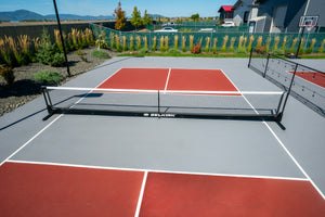 Outdoor pickleball court with low net labeled SELKIRK, red service boxes and gray playing surface.