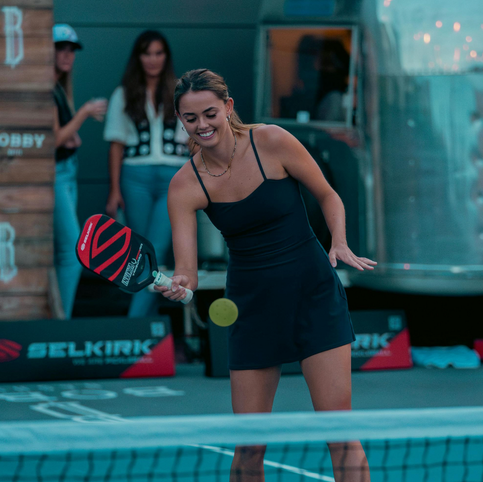A woman in a black dress playing pickleball with a paddle and ball.