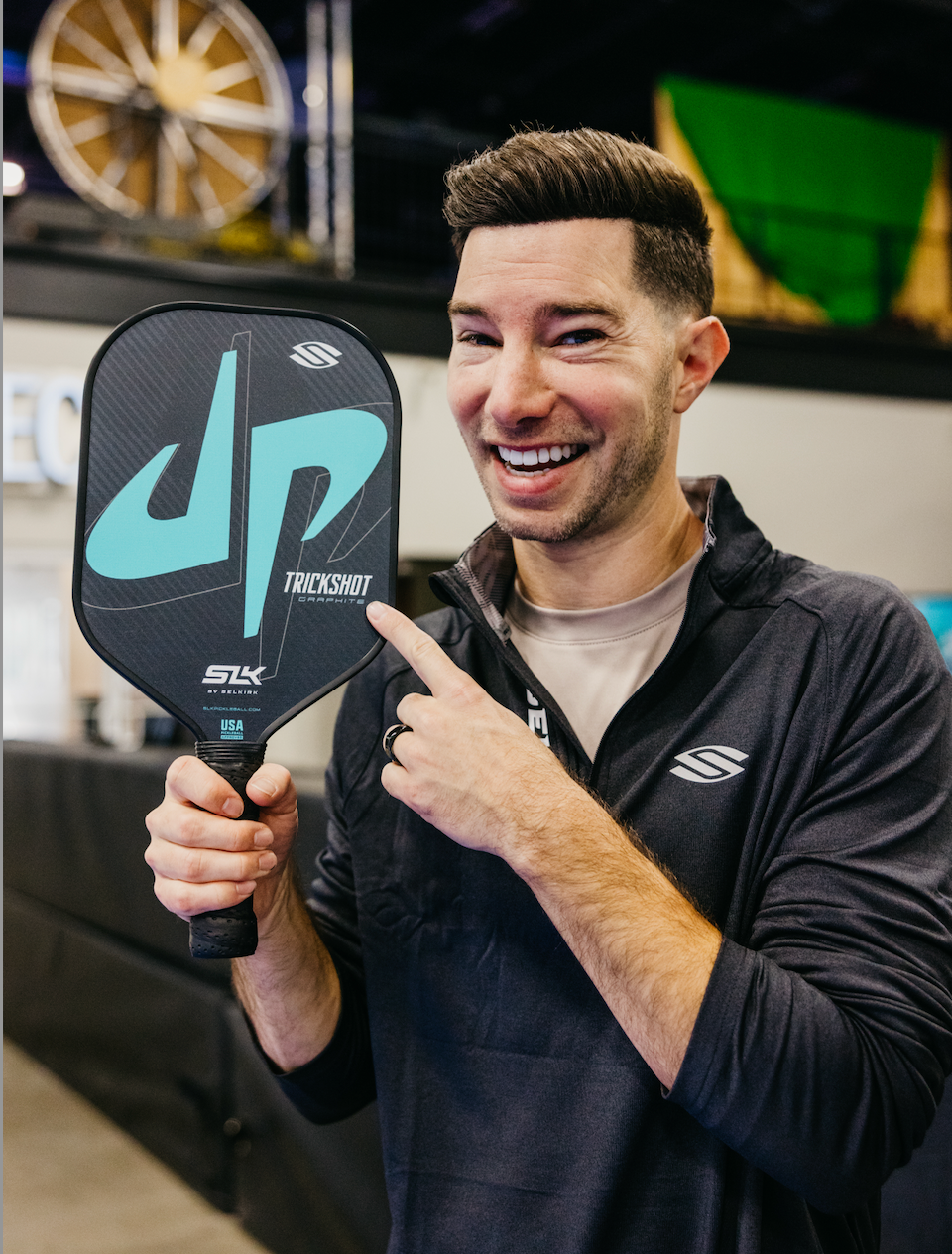 Cory Cotton holding the SLK by Selkirk x Dude Perfect Trickshot paddle, smiling and pointing at it.