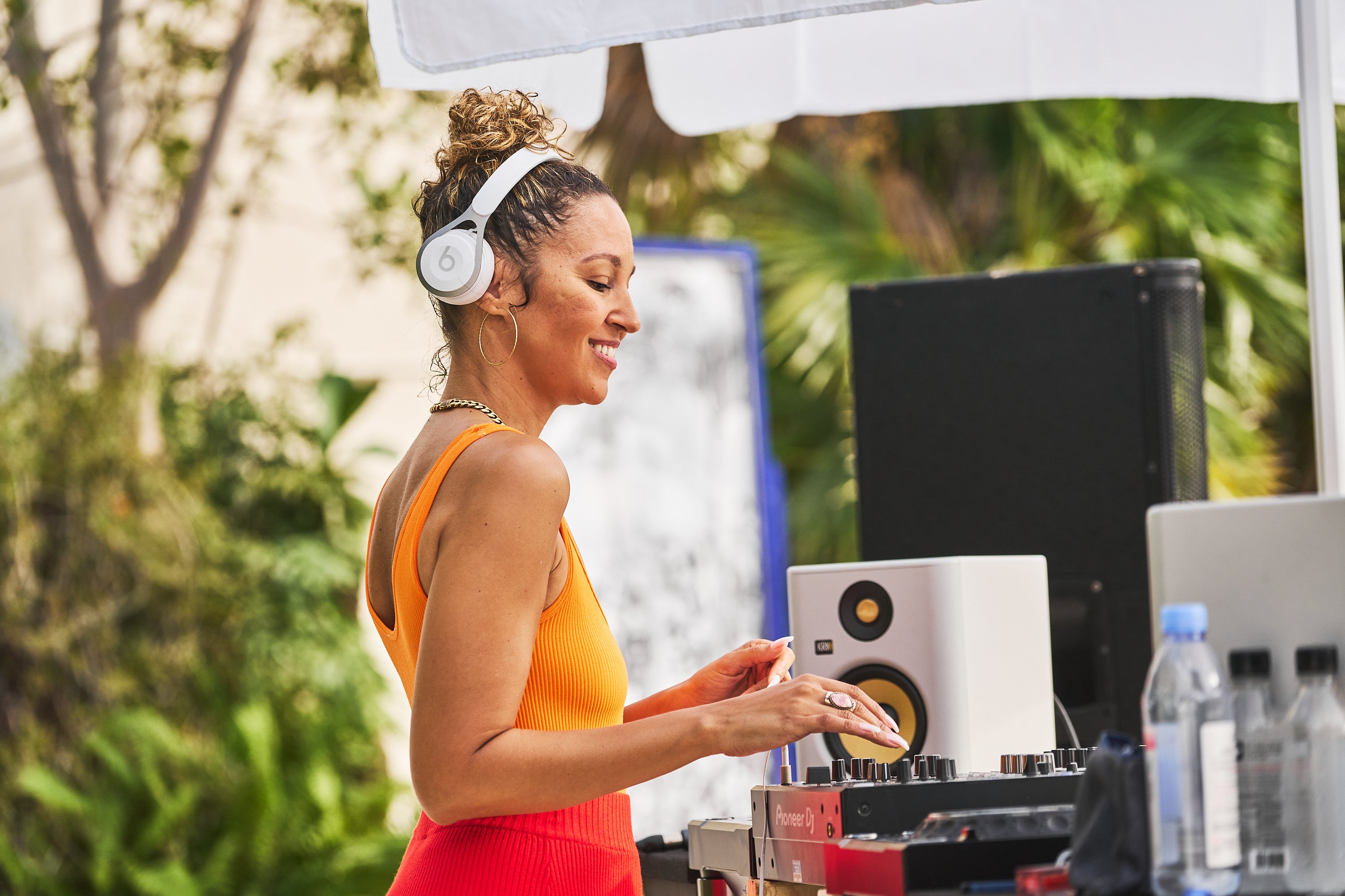 A woman in an orange dress DJing at an outdoor event with greenery in the background.
