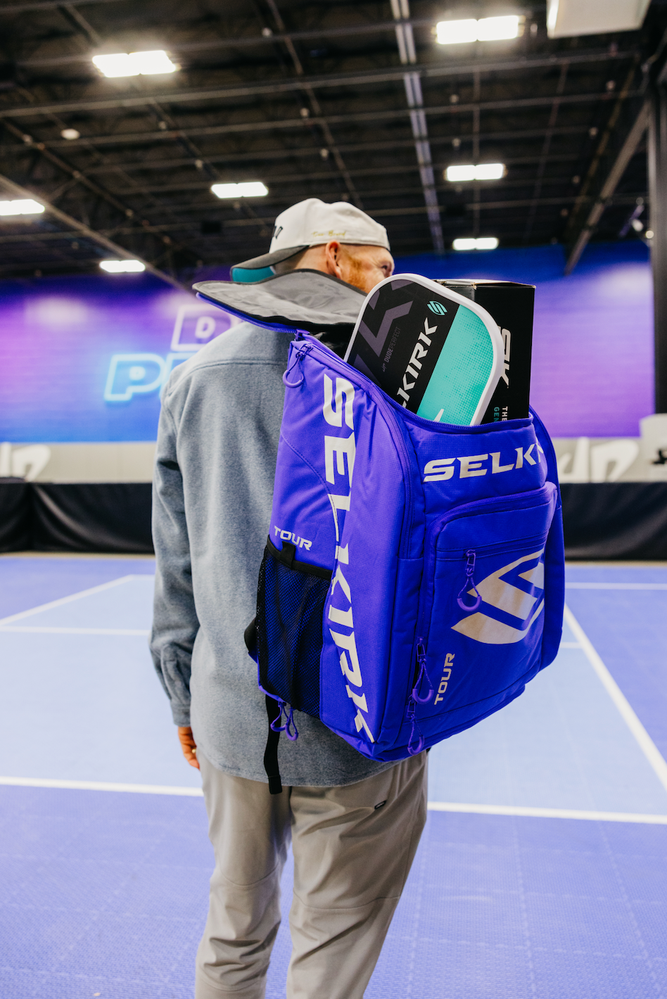 Person carrying a blue SELKIRK backpack with paddles on a pickleball court