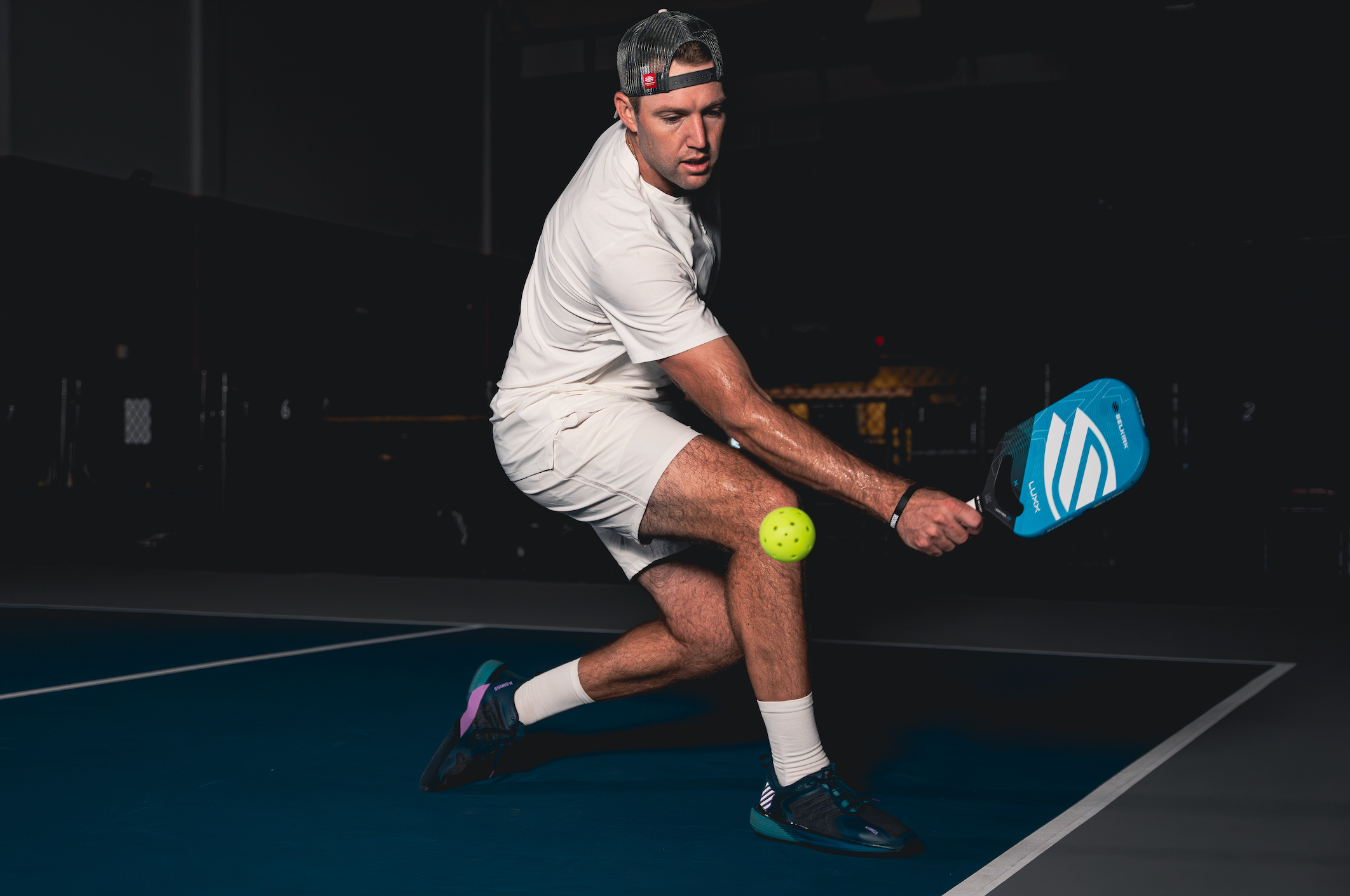 A male athlete in a white outfit hitting a pickleball with a blue paddle on a court.