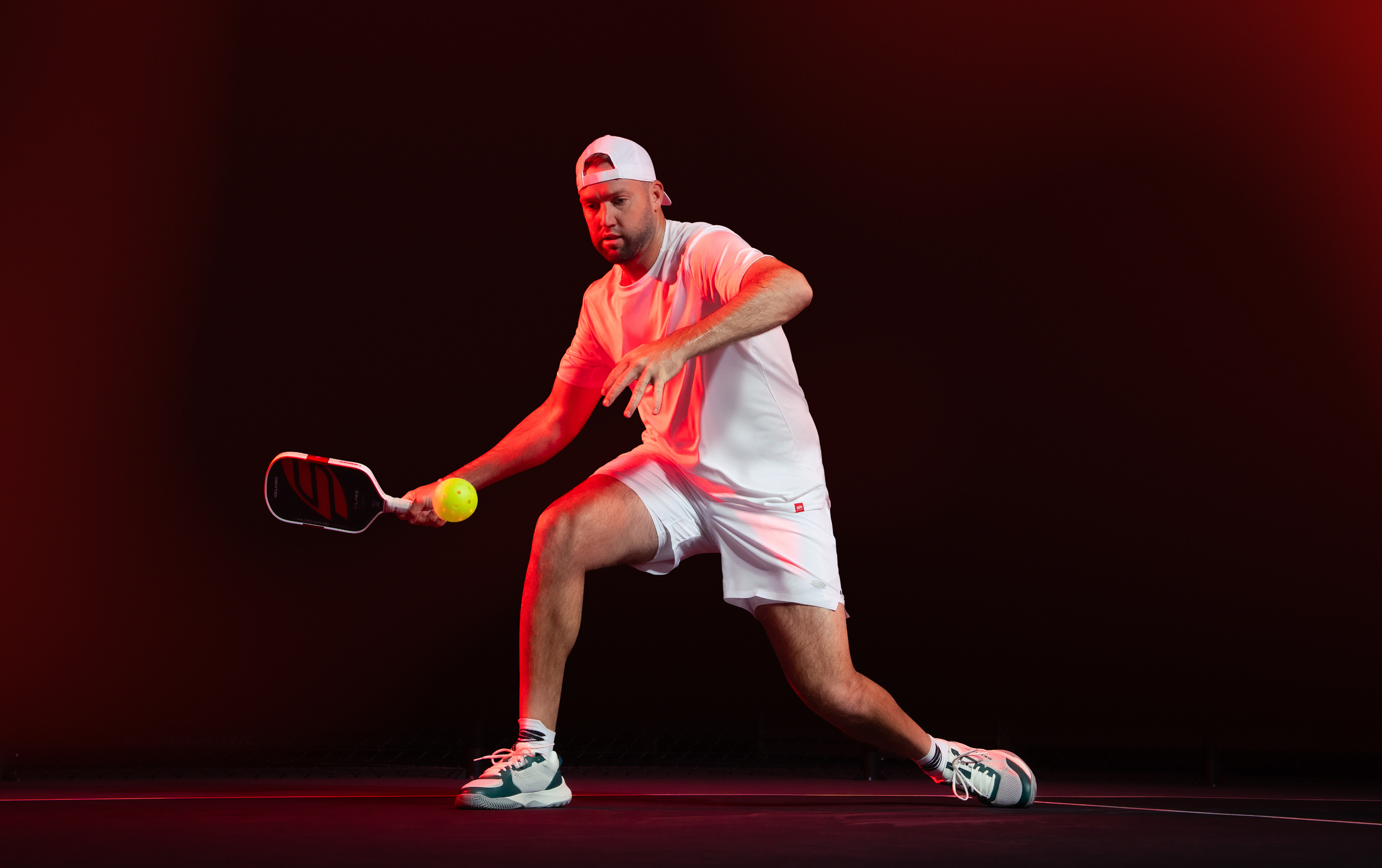 Male pickleball player lunging to volley a yellow ball with a paddle, wearing white athletic outfit and backward cap under red studio lighting.