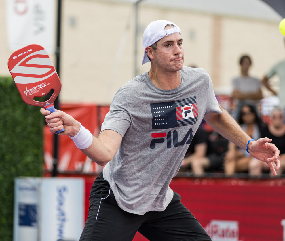 Male pickleball player swinging a red AMPED paddle on an outdoor court with spectators in the background