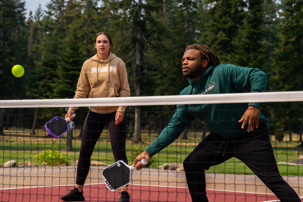 Two pickleball players at the net — man lunging to return a shot, woman standing ready, both wearing Selkirk hoodies.