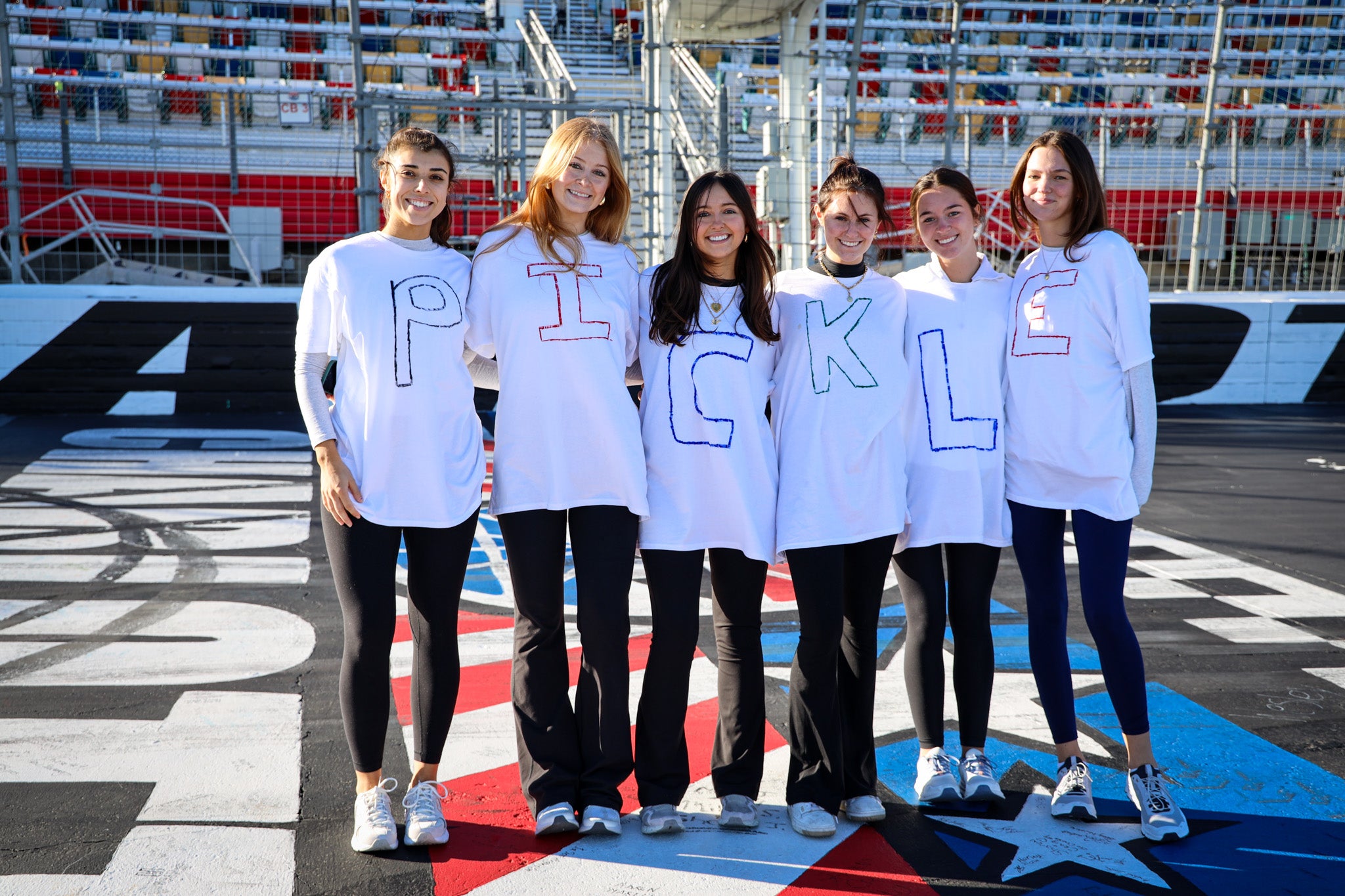 Six women wearing white shirts with letters spelling 'PICKLE' on a racetrack