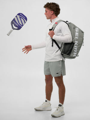 Young man in white long-sleeve and gray shorts tossing a purple pickleball paddle while carrying a gray SELKIRK backpack.