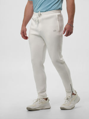 Men's off-white tapered jogger pants with drawstring waist, side pockets and ankle zippers, worn with cream sneakers.