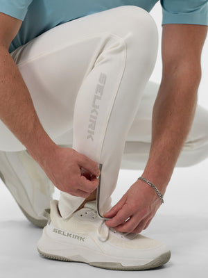 Kneeling person tightening zipper at ankle of white pants showing SELKIRK logo and white SELKIRK sneaker.