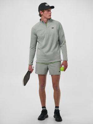 Male model in light-gray quarter-zip and shorts, black cap, holding a pickleball paddle and a green ball.