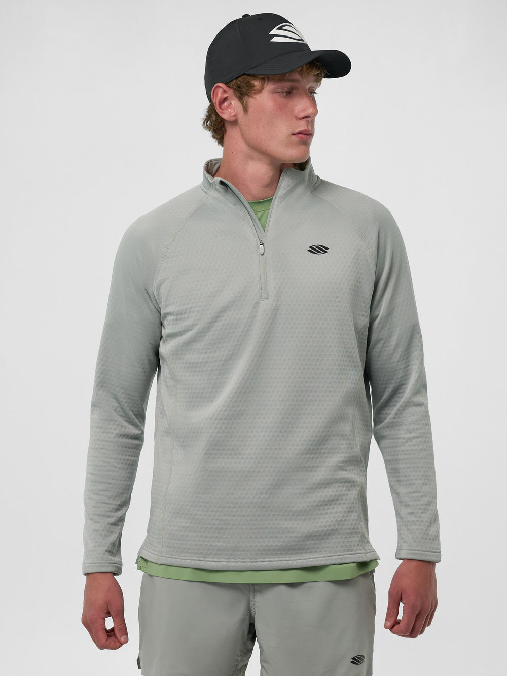 Model wearing a Selkirk men's quarter-zip shirt in light grey, with a black cap, standing against a neutral background.