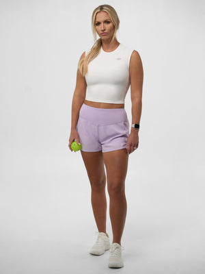 Female athlete wearing a white cropped top, lavender shorts and white sneakers, holding a small green ball