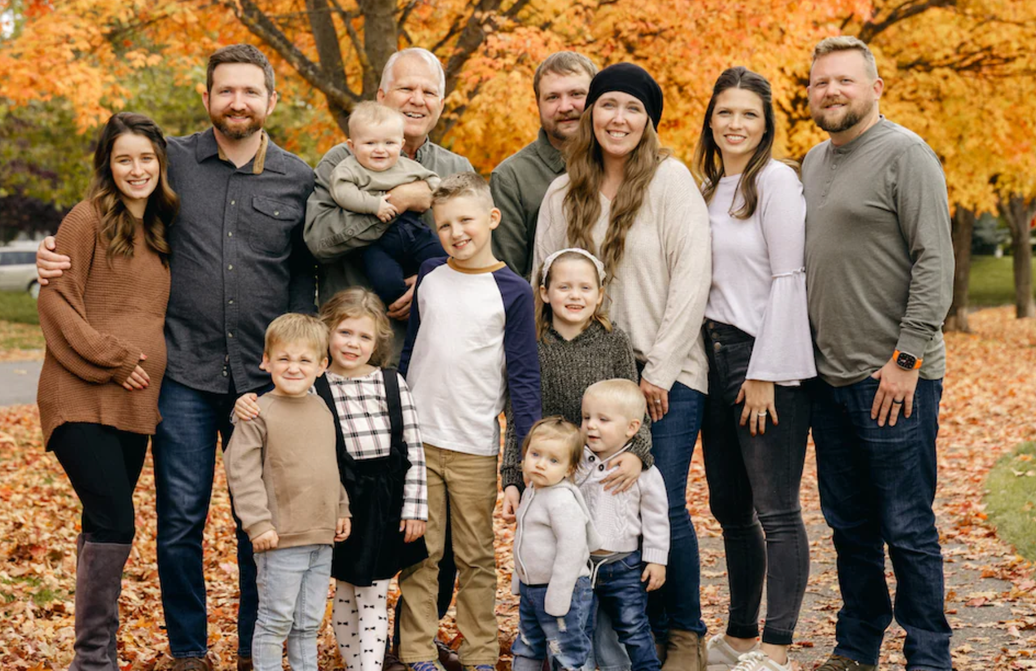 Large extended Selkirk family posed on a leaf-covered path with orange autumn trees, adults and children smiling together