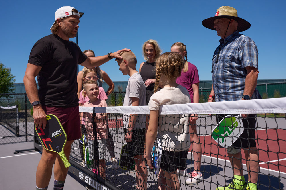 Coach pats a boy's head as children and two instructors stand at a pickleball net with paddles bearing SELKIRK and SLK logos.