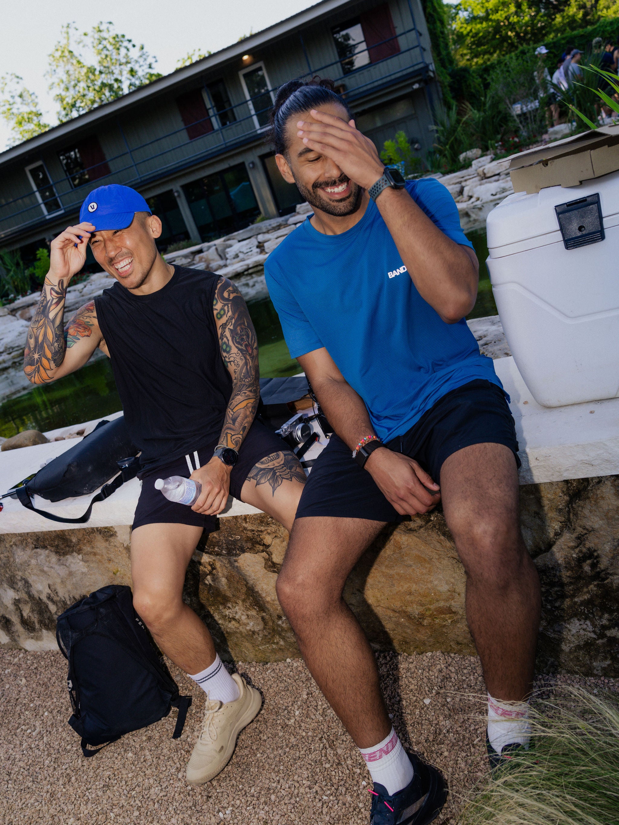 Two men smile and cover their eyes as they drink water next to a pickleball court. 