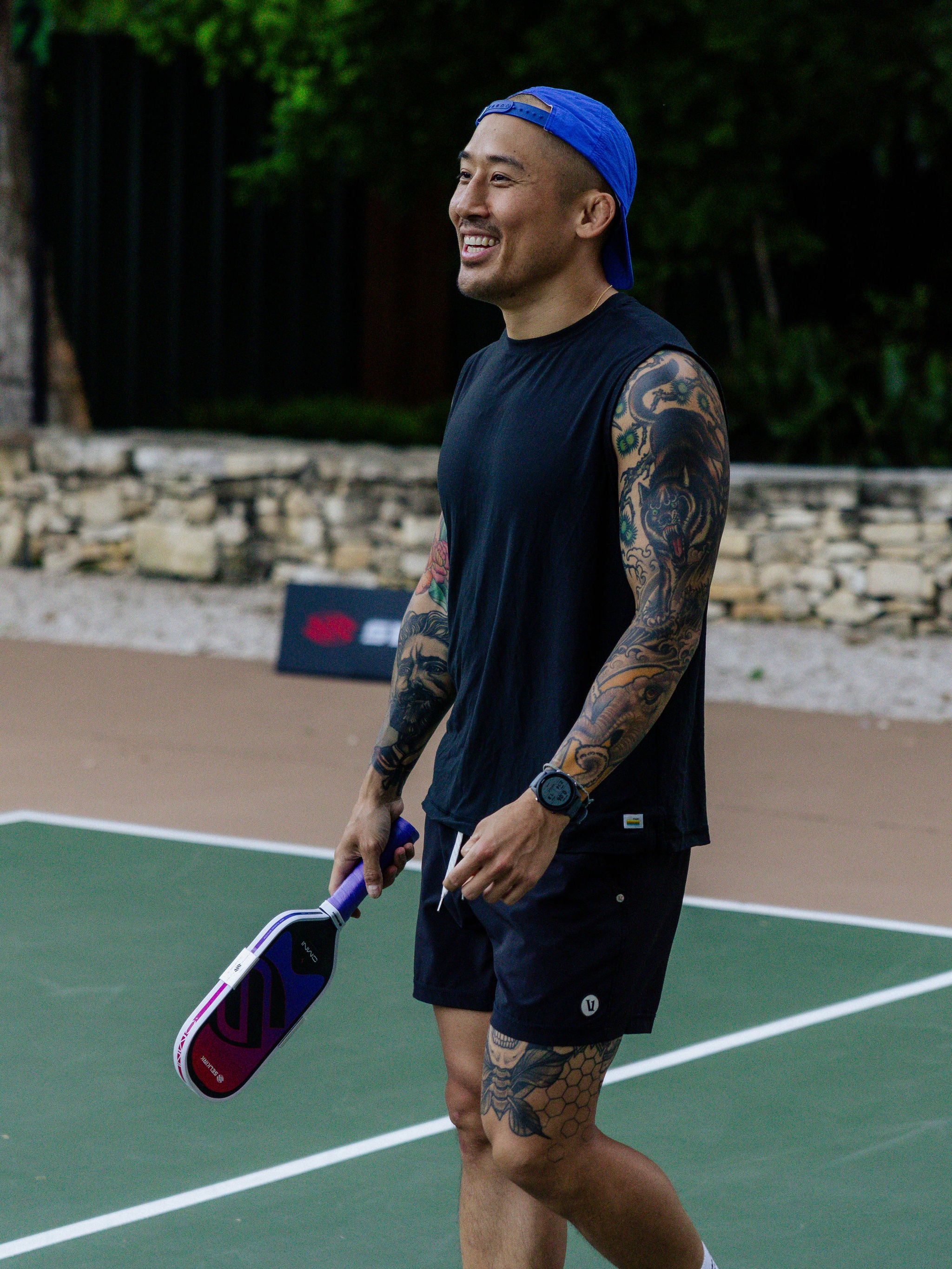 A man with tattoos smiles as he holds a Selkirk OMNI pickleball paddle on an outdoor pickleball court. 