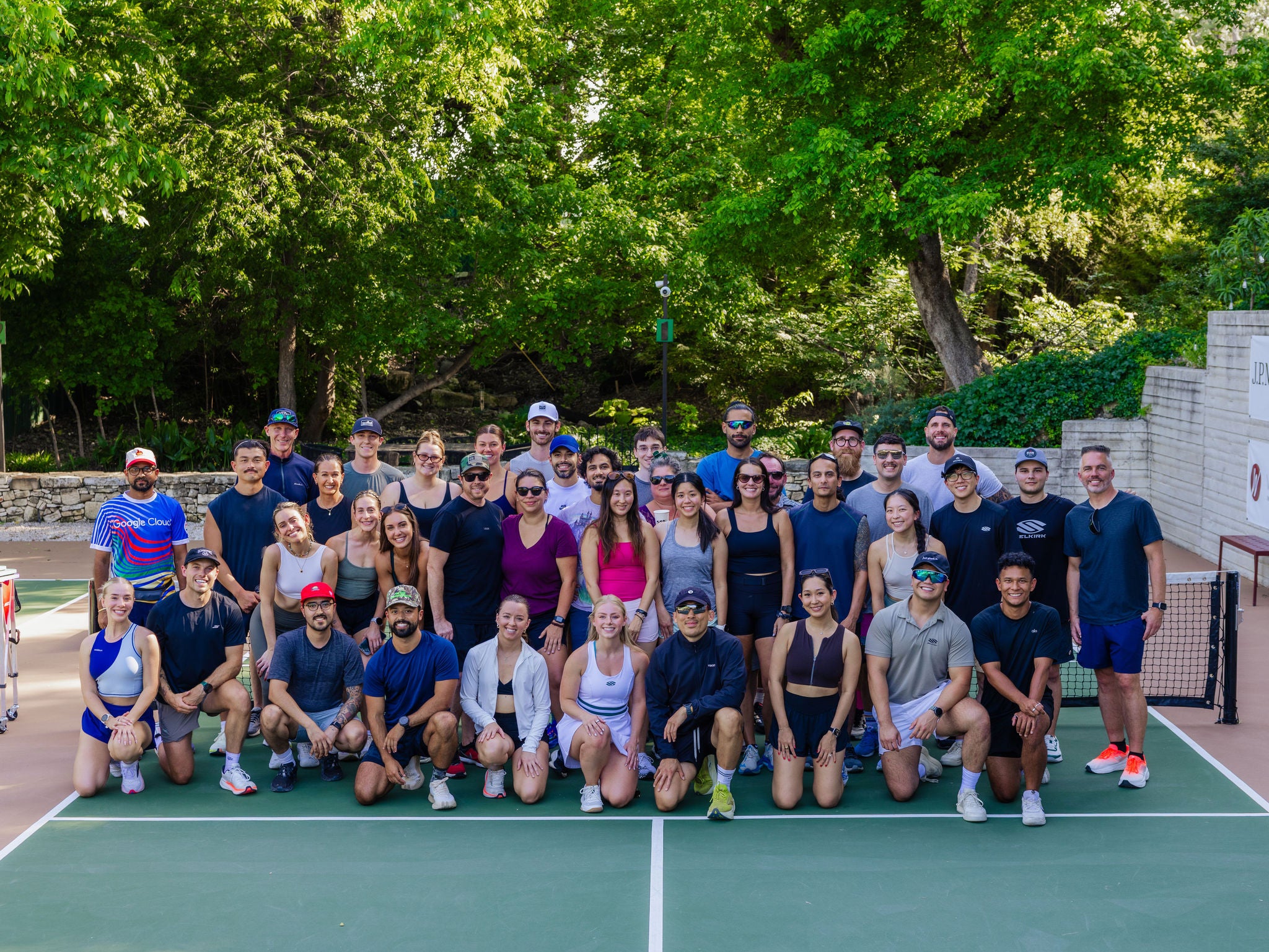 A large group of people stands in three lines and smiles for a camera on an outdoor pickleball court. 