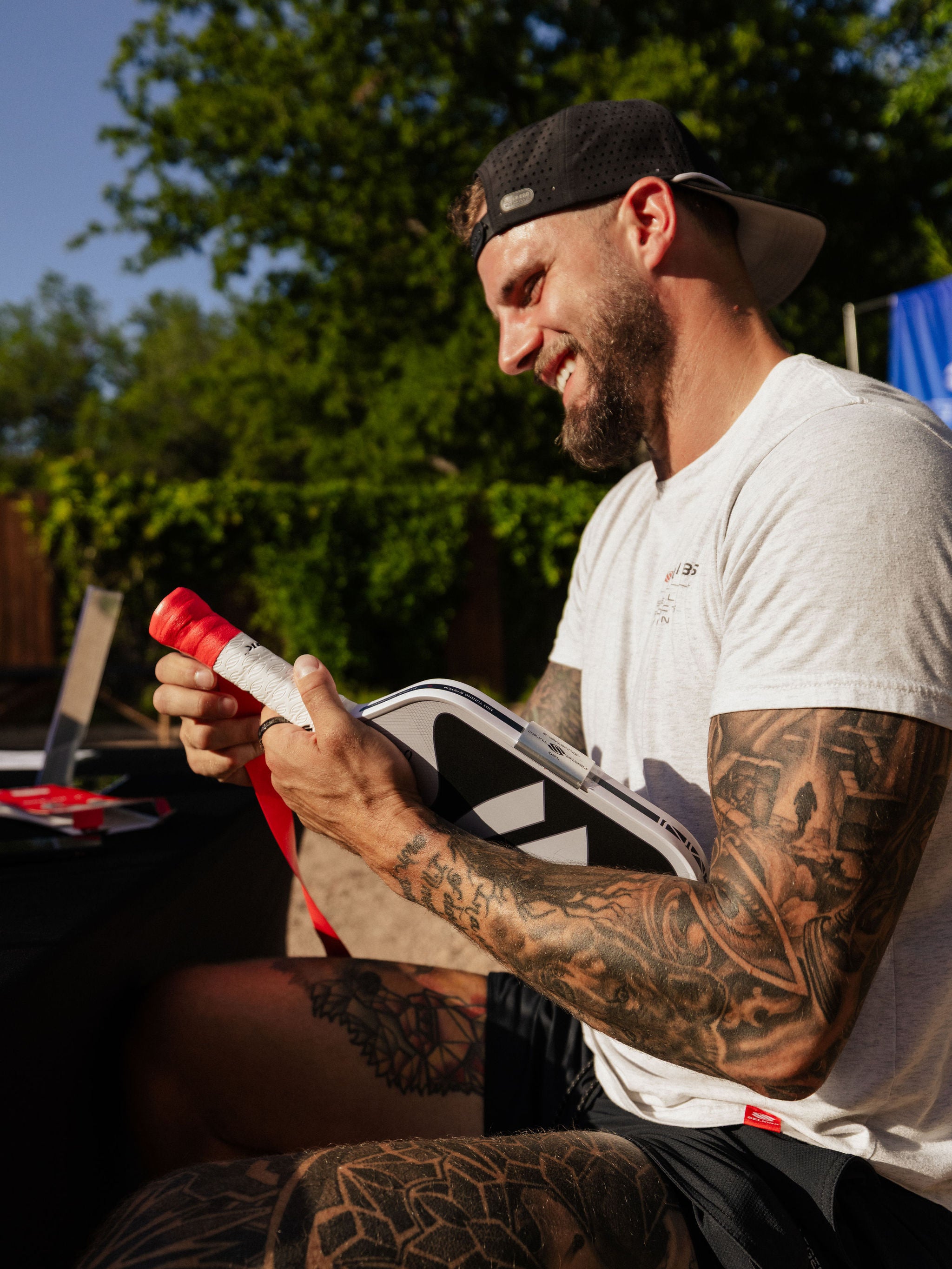 A man smiles as he wraps grip tape on a Selkirk pickleball paddle while seated. 
