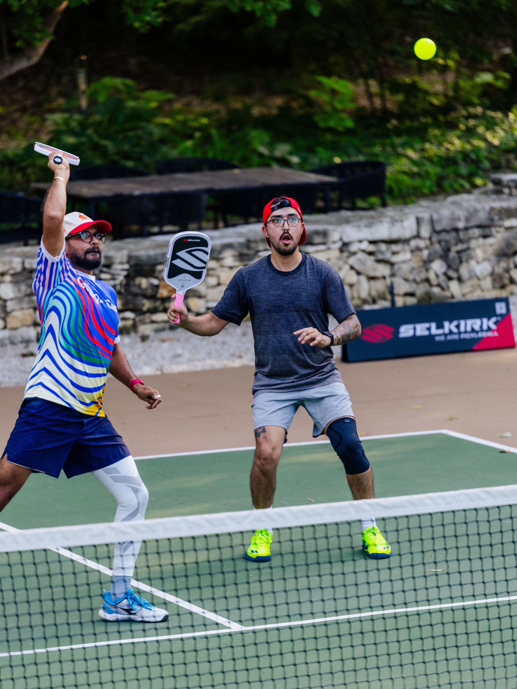 Two men raise their Selkirk OMNI paddles to hit an overhead shot on an outdoor pickleball court. 