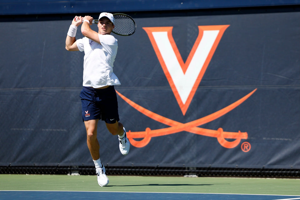 A University of Virginia athlete plays tennis. He is mid-hit in front of the UVA logo. 