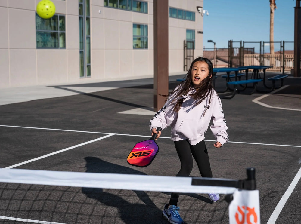 Young girl in a VANS hoodie playing pickleball on an outdoor court, swinging a colorful paddle toward a lime ball.
