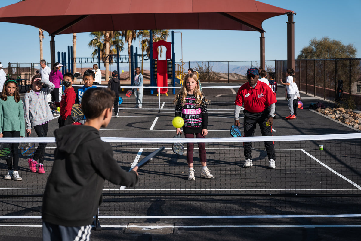 Children and instructor playing pickleball on outdoor courts under shade; girl wearing a sweater reading 'ON THE NAUGHTY LIST'.