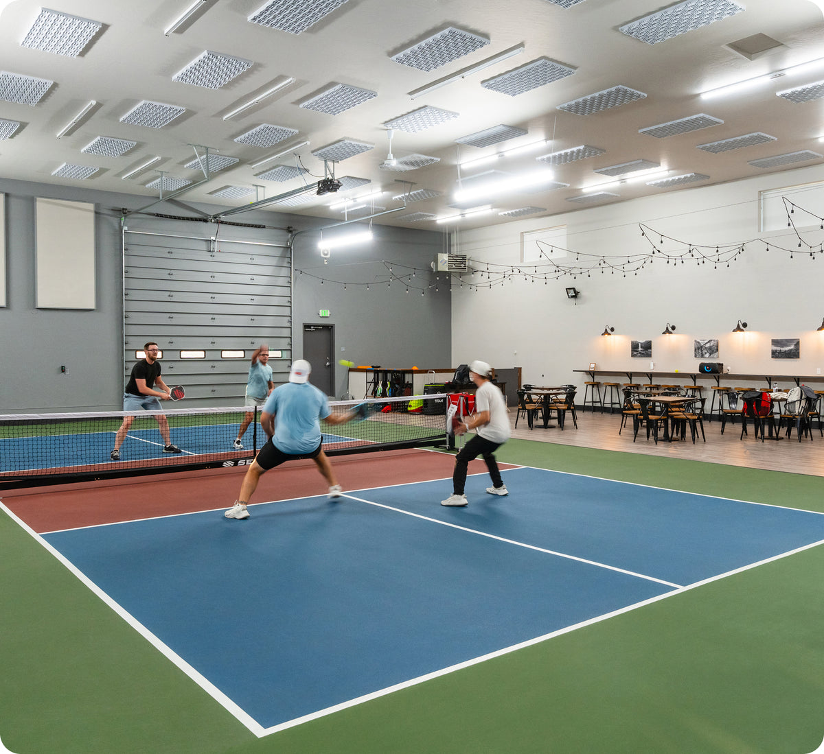 Indoor pickleball court with four players in a doubles match and a seating area with tables and string lights.