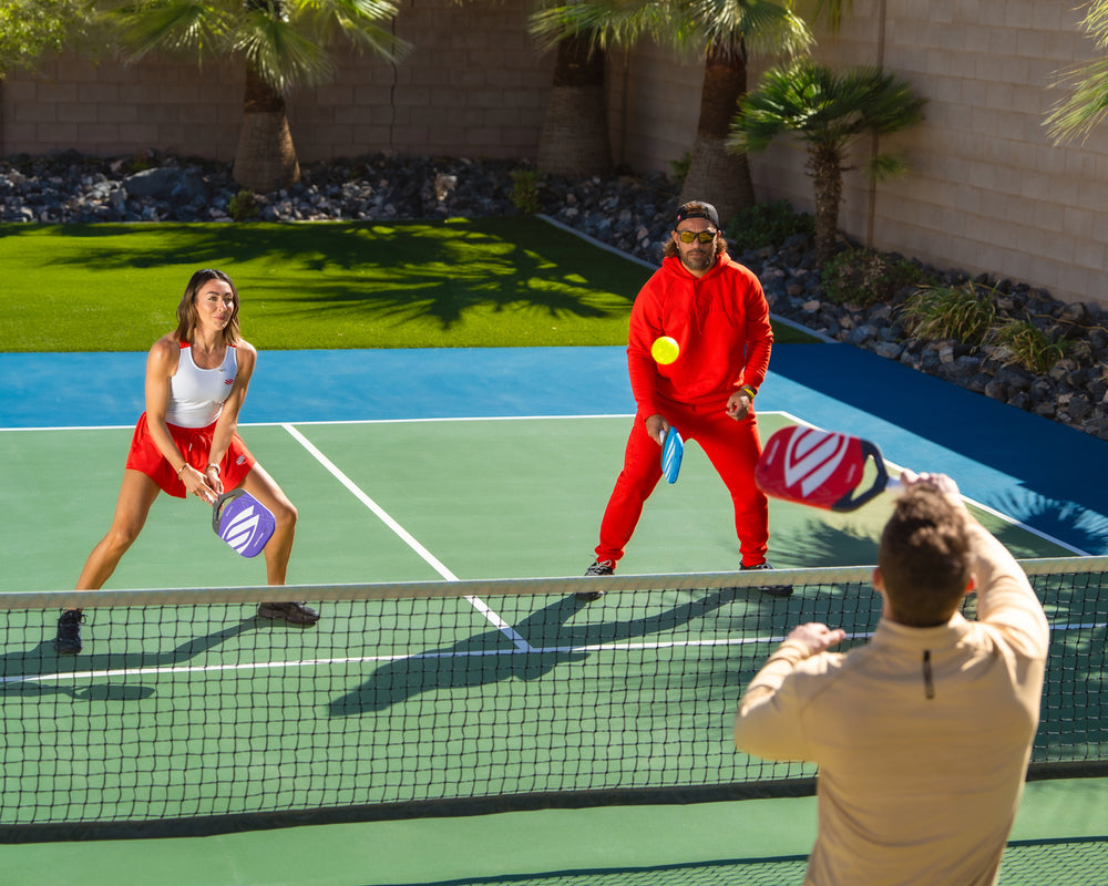 Three players in a backyard pickleball match: woman in white top and red skirt at net, man in red hoodie returning a lob, ball midair.