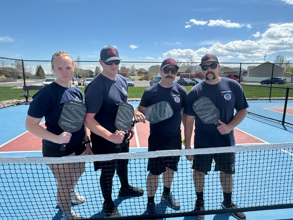 Four Lehi Fire firefighters holding pickleball paddles behind a net on an outdoor court on a sunny day.