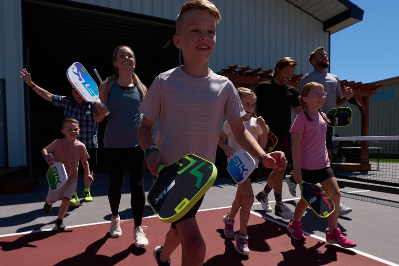 Group of children and adults carrying SLK pickleball paddles walking onto an outdoor court