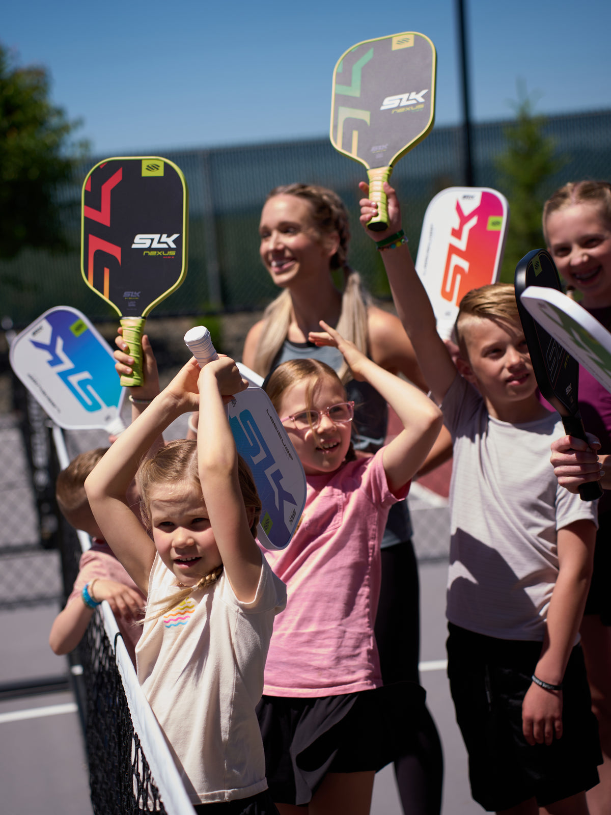 Children and an adult instructor raising Selkirk SLK pickleball paddles over a net on an outdoor court
