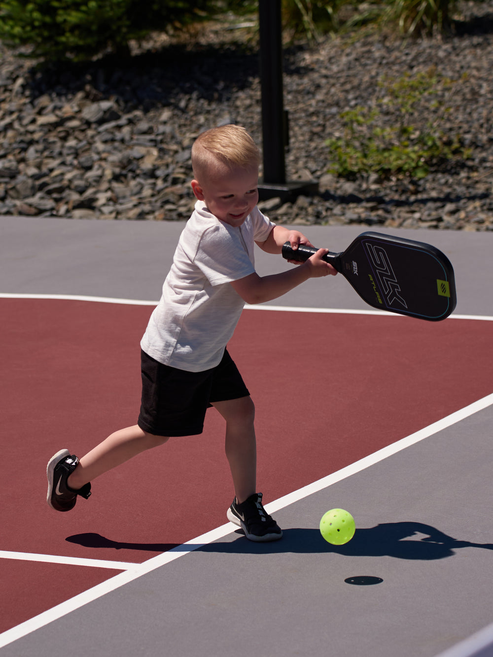 Young child swinging an SLK pickleball paddle to hit a neon ball on an outdoor court