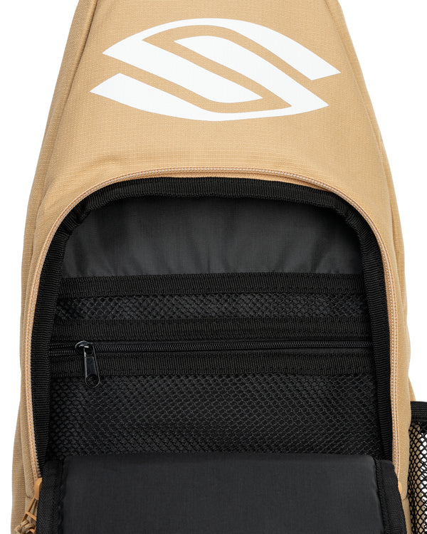 Tan sling backpack opened, showing interior black padded compartment, mesh zip pocket, and white stylized logo on flap.