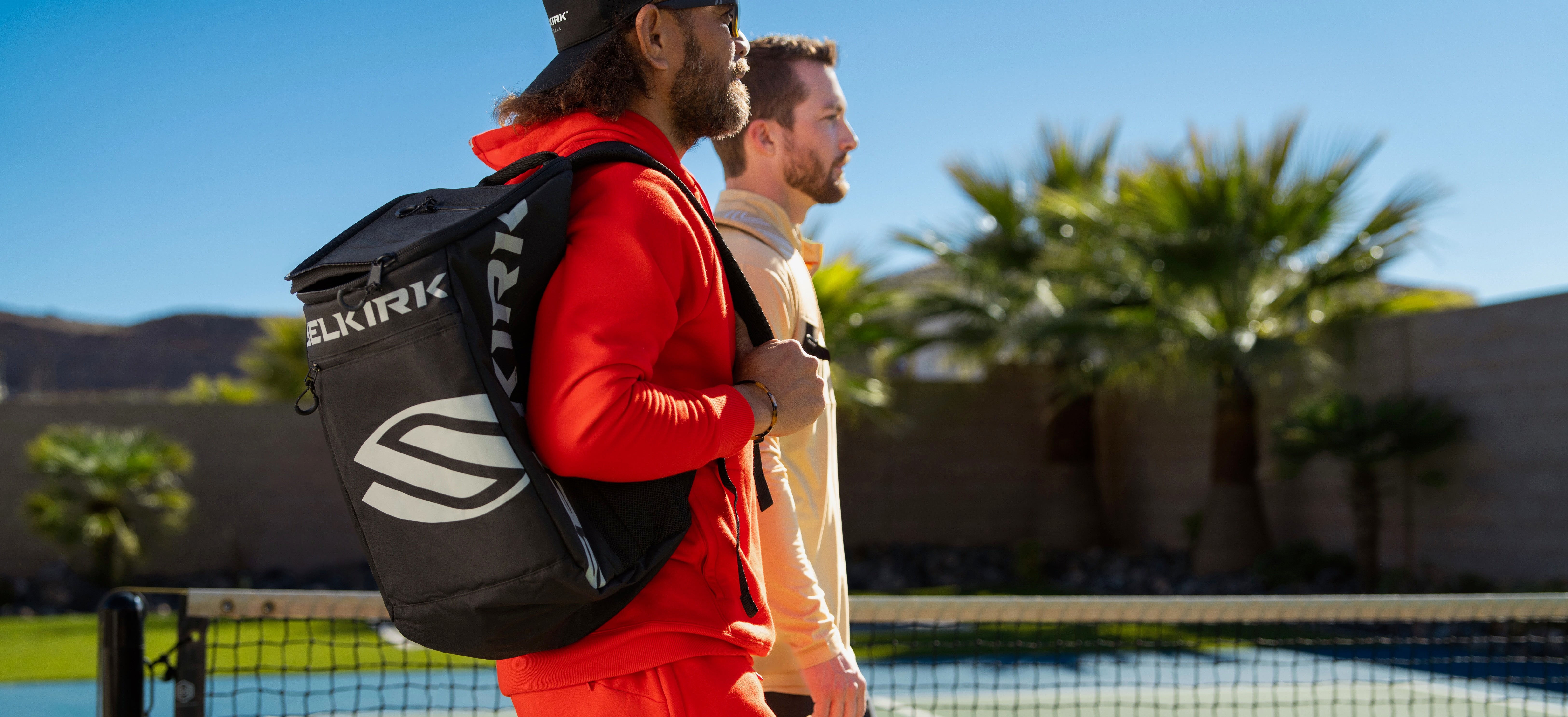 Side view of Selkirk pickleball backpack worn by a player, showing large Selkirk logo and padded shoulder straps with a sunny outdoor pickleball court in the background