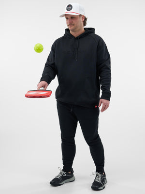 Man wearing black Selkirk hoodie and cap holding a red Selkirk pickleball paddle, bouncing a yellow pickleball.