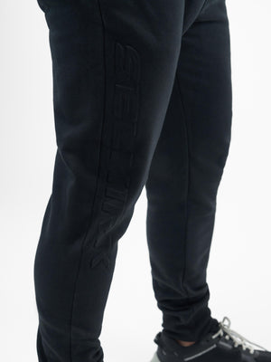 Close-up of lower legs wearing black athletic joggers with a vertical embossed logo on the left calf and black sneakers.