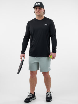 Man in black long-sleeve shirt and black cap holding pickleball paddle and neon yellow pickleball, wearing light green shorts