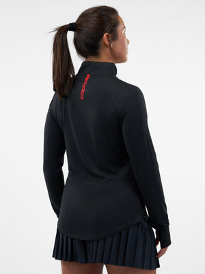 Woman seen from back wearing black long-sleeve top with red vertical logo at collar and pleated black skirt, hair in ponytail