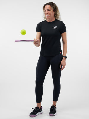 Woman wearing black workout top and leggings smiling while balancing a green ball on a purple paddle