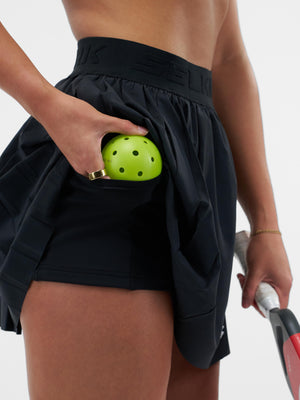 Player placing neon green perforated pickleball into the pocket of a black skort, paddle held in the other hand.
