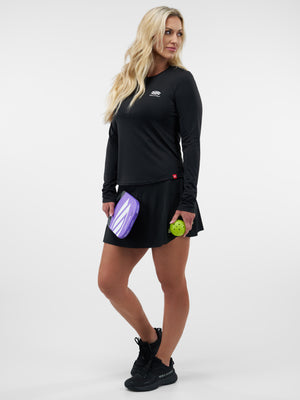 Woman in black athletic top and skirt holding a purple pickleball paddle and a green pickleball, looking to her right.