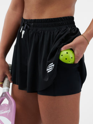 Black athletic skort with built-in shorts; hand holding neon green perforated pickleball in pocket, pink paddle at side